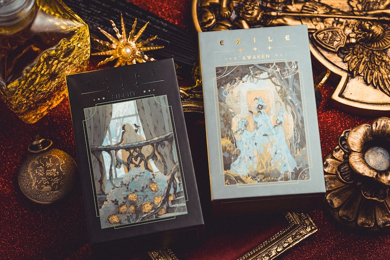 Op de afbeelding: Twee tarotkaartenboxen met de woorden "EXILE" en "AWAKEN" in gouden letters. De dozen bevatten illustraties van figuren in een fantasiewereld. De achtergrond is een rood glitteroppervlak met gouden decoratieve elementen.
