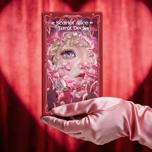 Puede incluir: Una caja rectangular de baraja de tarot Scarlet Alice con una ilustración detallada de un rostro rodeado de conejos rosas y tazas de té florales. La caja tiene un borde rojo intenso con detalles ornamentales en blanco.