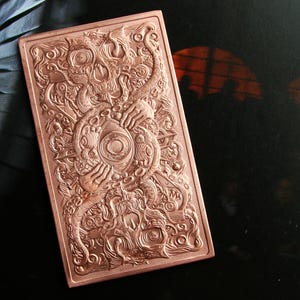 May include: A rectangular copper-colored metal bar featuring an intricate, embossed design. The artwork displays a central eye surrounded by tentacles, hands, and swirling patterns, creating a highly detailed, textured surface across the entire face.