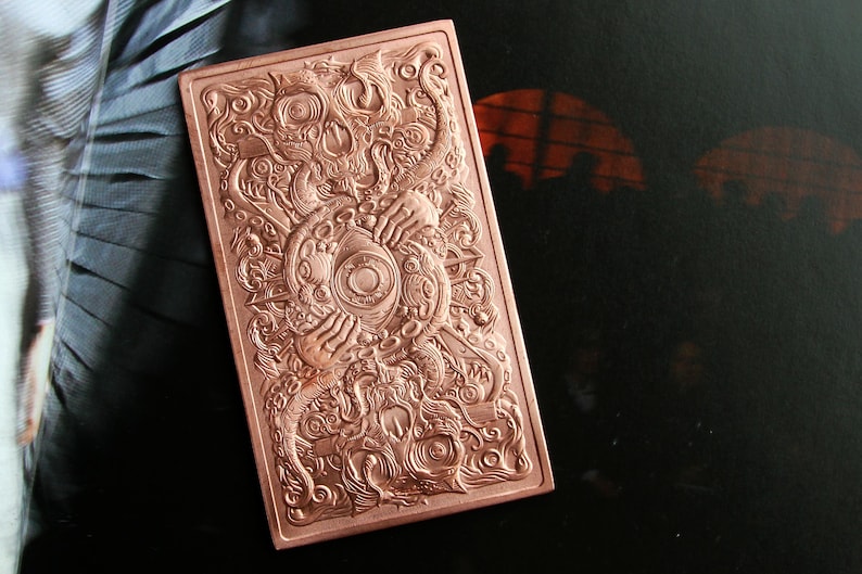 May include: A rectangular copper-colored metal bar featuring an intricate, embossed design. The artwork displays a central eye surrounded by tentacles, hands, and swirling patterns, creating a highly detailed, textured surface across the entire face.