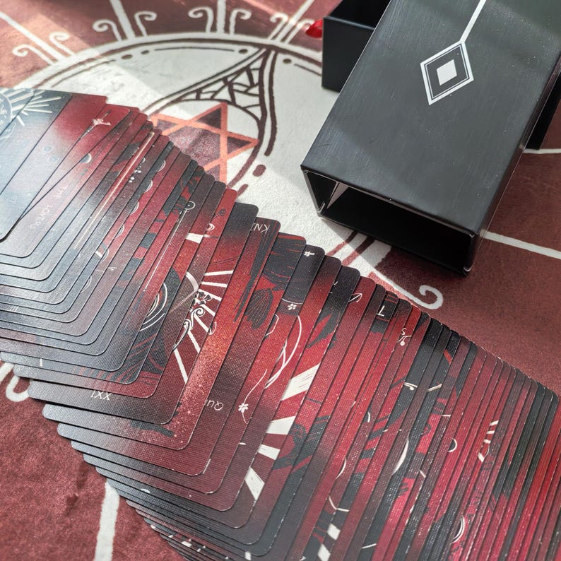 Puede incluir: Una baraja de cartas de tarot con un dise&ntilde;o rojo y negro, extendida sobre una superficie estampada. Las cartas presentan ilustraciones y texto en blanco. Se ve una caja negra con un motivo de diamante.