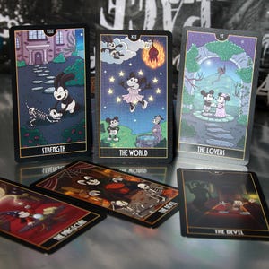 May include: A tarot card deck featuring cartoon characters. The cards have black borders and gold accents. The cards include the Strength, The World, The Lovers, The Magician, and The Devil cards. The illustrations are colorful and detailed.