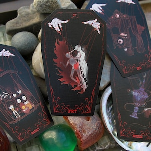 May include: Four coffin-shaped cards with black backgrounds and red line art illustrations. The cards feature marionette hands, a figure labeled Spirit, a market scene, and an incense burner. Each card has a unique number and text at the bottom.