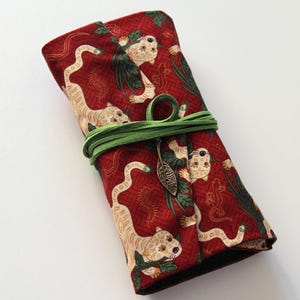 May include: A rolled fabric organiser featuring a deep red background with a repeating pattern of cream-coloured tigers and green foliage. The roll is secured with a long, thin green cord that wraps around the centre and features a metal leaf charm.