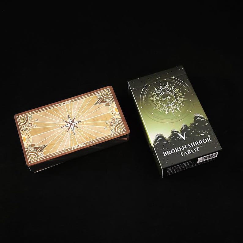 May include: A deck of Broken Mirror Tarot cards featuring a tan, star-patterned back design and a green box with a sun illustration, the Roman numeral V, and the text Broken Mirror Tarot printed in white on the front of the packaging.