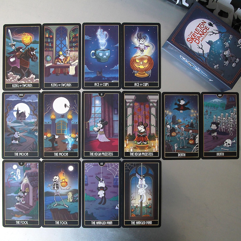 May include: A deck of tarot cards with a Halloween theme. Each card features a unique illustration with characters and symbols. The box reads "The Skeleton Dance Arcana". The cards are displayed on a light gray surface.