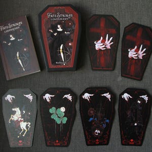 May include: A deck of Fate Strings Lenormand cards featuring coffin-shaped cards with dark illustrations of marionette hands, a white horse, clover, and a ship. The set includes a rectangular box and a coffin-shaped box with matching artwork.