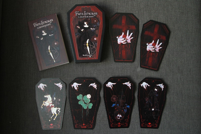 May include: A deck of Fate Strings Lenormand cards featuring coffin-shaped cards with dark illustrations of marionette hands, a white horse, clover, and a ship. The set includes a rectangular box and a coffin-shaped box with matching artwork.
