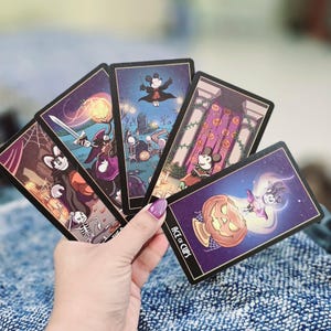 May include: A hand holding a fanned deck of Halloween-themed tarot cards. Each card features a different illustration with a spooky theme, including a skeleton, a pumpkin, and a bat. The Ace of Cups card is visible.