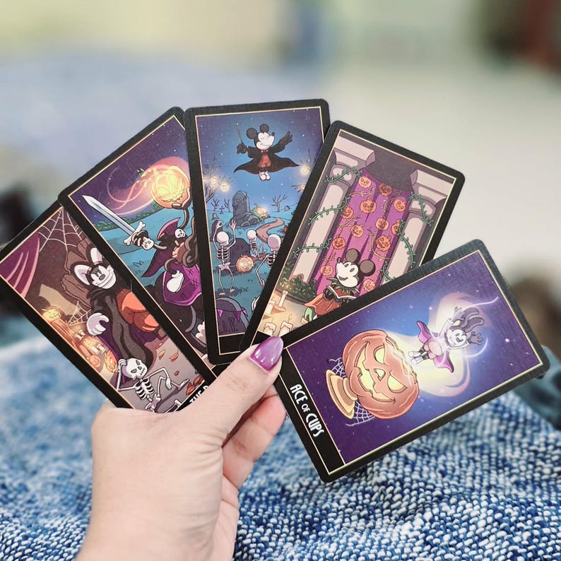 May include: A hand holding a fanned deck of Halloween-themed tarot cards. Each card features a different illustration with a spooky theme, including a skeleton, a pumpkin, and a bat. The Ace of Cups card is visible.