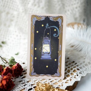 May include: A vintage-style tarot card with a gold frame and a dark blue lantern with a glowing light inside. The lantern is surrounded by small yellow stars on a dark blue background.
