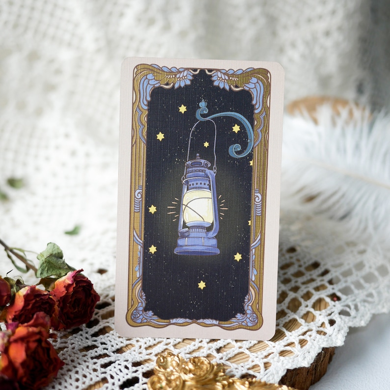 May include: A vintage-style tarot card with a gold frame and a dark blue lantern with a glowing light inside. The lantern is surrounded by small yellow stars on a dark blue background.