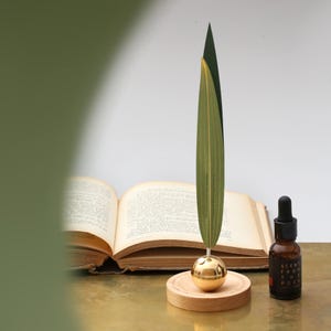 May include: A decorative leaf-shaped ornament with a gold sphere base, on a wooden stand. An open book and a small brown bottle with a dropper are in the background. The bottle has text that reads "SCENT FROM THE POEM".