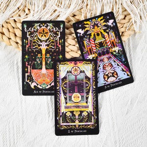 May include: Three tarot cards with intricate illustrations are displayed. The cards feature vibrant colors and detailed artwork, including images of a tree, a window, and a celestial design. The cards are labeled "Ace of Pentacles", "2 of Pentacles", and "3 of Pentacles".