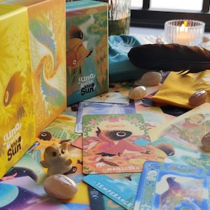May include: A collection of tarot cards, crystals, and decorative items. The image features two boxes labeled "Uno, Beneath the Sun", a blue pouch, a black feather, and several cards with illustrations. The color palette includes yellow, blue, and earth tones.