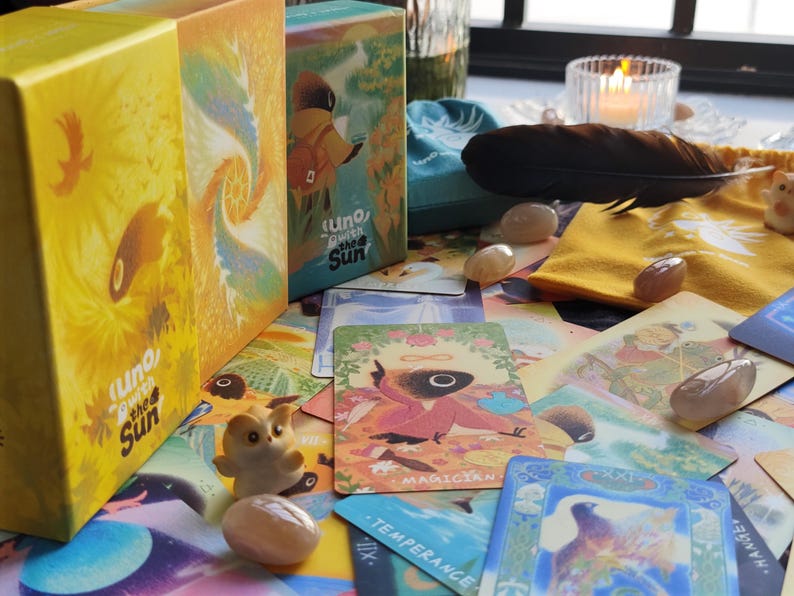 May include: A collection of tarot cards, crystals, and decorative items. The image features two boxes labeled "Uno, Beneath the Sun", a blue pouch, a black feather, and several cards with illustrations. The color palette includes yellow, blue, and earth tones.