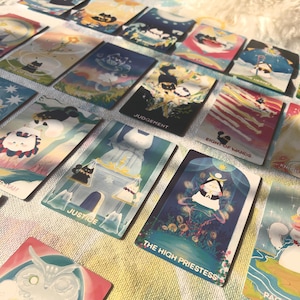 K&ouml;nnte beinhalten: Eine Sammlung rechteckiger Tarotkarten mit farbenfrohen Illustrationen von Katzen in verschiedenen Posen und Umgebungen. Sichtbarer Text umfasst The High Priestess, Justice, Judgement, Eight of Wands und Ace of Cups auf den Kartenoberfl&auml;chen.