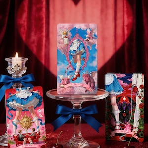 May include: Three rectangular tarot cards featuring whimsical illustrations of a character in a blue dress and red shoes. The center card is labeled 21. The World, the left card 19. The Sun, and the right card 15. The Devil, all displayed on a glass stand.