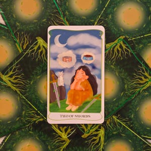 May include: A tarot card illustration featuring a woman in an orange dress dreaming of cat food, with a white cat and two swords. The card is labeled "TWO OF SWORDS" and set against a background of green foliage.
