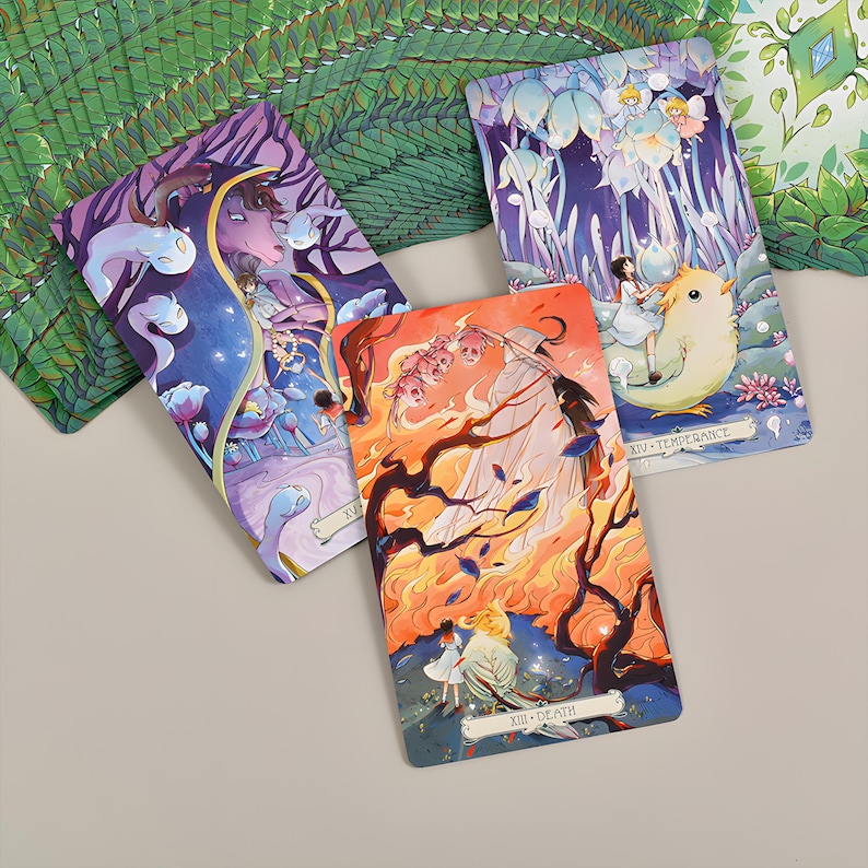 May include: Three tarot cards with colorful illustrations. One card features a figure with a bird, another has a figure with a creature, and the third depicts a figure with a creature in a fiery scene. The cards are on a light gray surface.