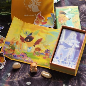 May include: A collection of illustrated items, including a yellow Uno bag, stickers, and a card game box. The box contains cards with a blue and white illustration. The scene is set on a dark, patterned surface with small decorative stones.