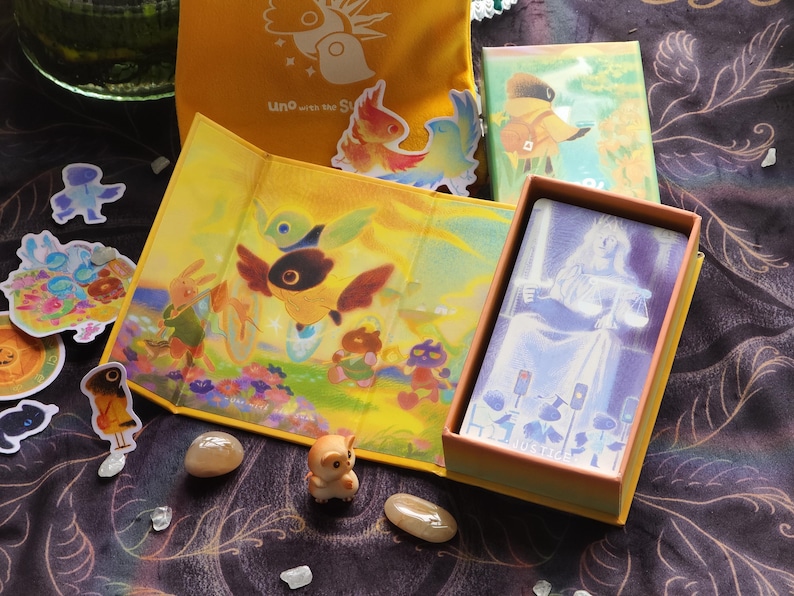 May include: A collection of illustrated items, including a yellow Uno bag, stickers, and a card game box. The box contains cards with a blue and white illustration. The scene is set on a dark, patterned surface with small decorative stones.