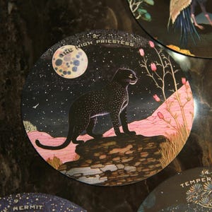 May include: Circular tarot card featuring a black panther under a starry night sky with a full moon. The card is labeled "THE HIGH PRIESTESS" in white text. Pink and green flora are also present.