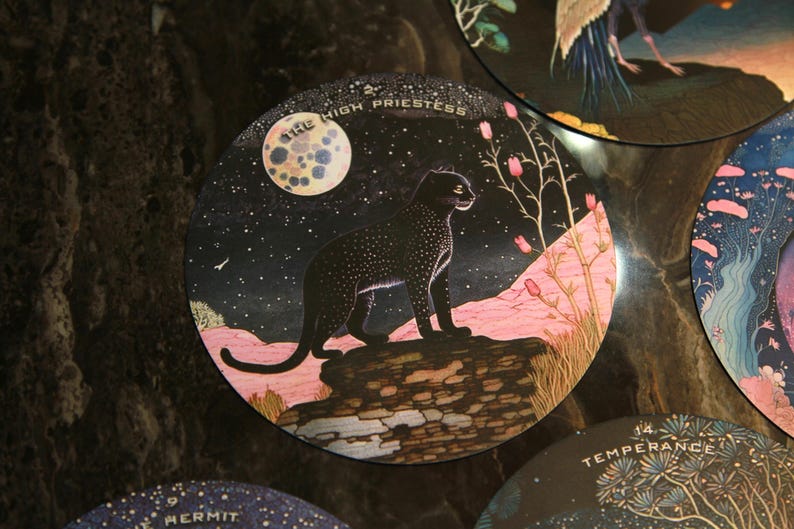 May include: Circular tarot card featuring a black panther under a starry night sky with a full moon. The card is labeled "THE HIGH PRIESTESS" in white text. Pink and green flora are also present.