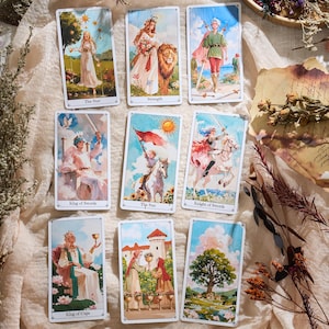 May include: A collection of tarot cards featuring colorful illustrations. The cards depict figures in various scenes, including a woman with a lion, a figure with a sword, and a person holding a cup. The cards have white borders and text labels.