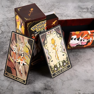May include: A tarot card deck with a brown box featuring the text "FANTASY THE Tarot". The cards have gold edges and illustrations of figures and floral designs. One card reads "THREE OF WANDS", another reads "ACE OF WANDS".