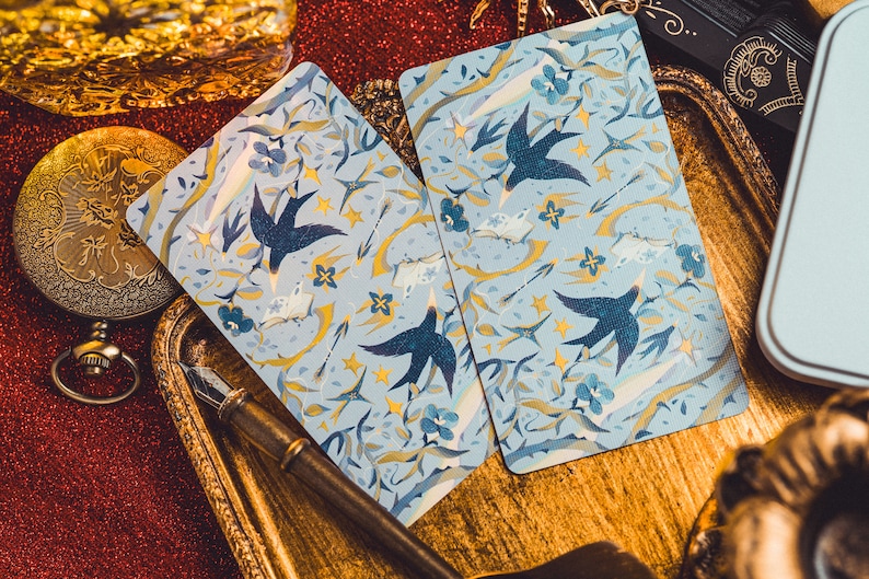 Op de afbeelding: Twee speelkaarten met een lichtblauwe achtergrond met een patroon van donkerblauwe vogels, gele sterren en bloemelementen. De kaarten liggen op een gouden dienblad met een vintage pen en een zakhorloge.