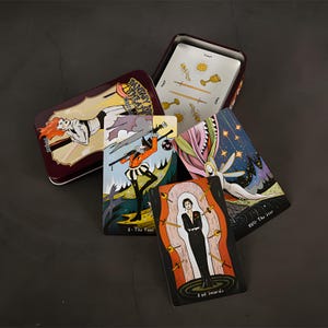 May include: A maroon metal tin with a tarot card deck. The cards feature colorful illustrations of figures and symbols. The tin has the words "Fantasy Art Theatre" on the lid. Some cards are fanned out, displaying various designs.