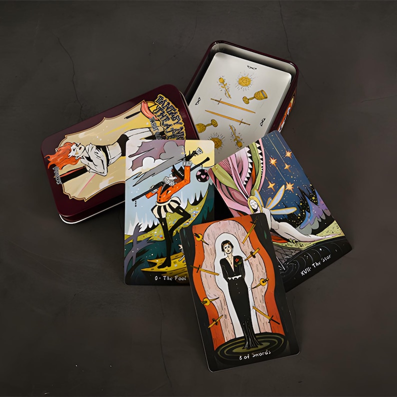 May include: A maroon metal tin with a tarot card deck. The cards feature colorful illustrations of figures and symbols. The tin has the words "Fantasy Art Theatre" on the lid. Some cards are fanned out, displaying various designs.