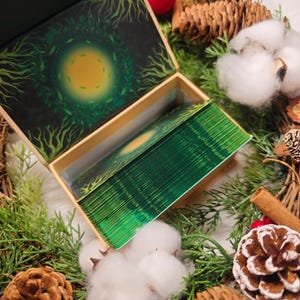 May include: A deck of tarot cards in a decorative box. The box and cards feature a green and gold design with a central sun-like image surrounded by a leafy border. The cards are green-edged and the box is open, revealing the deck.
