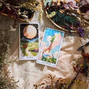 May include: Two tarot cards, "The Moon" and "Ace of Wands", are displayed on a beige cloth, surrounded by dried flowers and decorative elements. The Moon card features a fox and a path under a large moon. The Ace of Wands card shows a hand holding a wand.