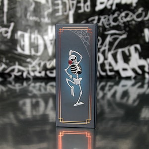 May include: A dark blue rectangular box featuring a dancing skeleton with a red rose in its mouth and a spider web. The box has gold-colored trim and a gradient blue background. The design is a whimsical take on the Day of the Dead theme.