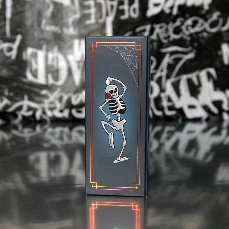May include: A dark blue rectangular box featuring a dancing skeleton with a red rose in its mouth and a spider web. The box has gold-colored trim and a gradient blue background. The design is a whimsical take on the Day of the Dead theme.