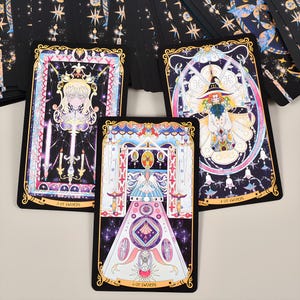 May include: A set of tarot cards featuring colorful illustrations with gold accents. The cards depict figures and symbols within ornate frames, with titles like "3 of Swords" and "4 of Swords" visible. The overall design has a mystical theme.