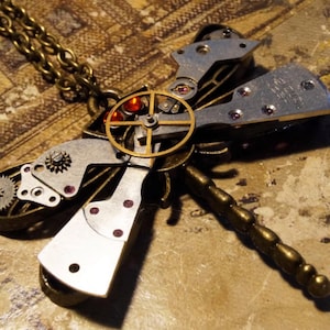 May include: A bronze and silver steampunk dragonfly necklace with a clockwork design and red and purple rhinestones. The necklace has a chain and a dragonfly pendant.
