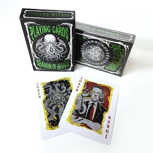 May include: Black playing card boxes with green and white text and illustrations. The boxes read "Cthulhu Mythos Playing Cards Shadow of Aeons" and "Art by Doubleem". Two joker cards are visible, featuring illustrations and the word "Joker".