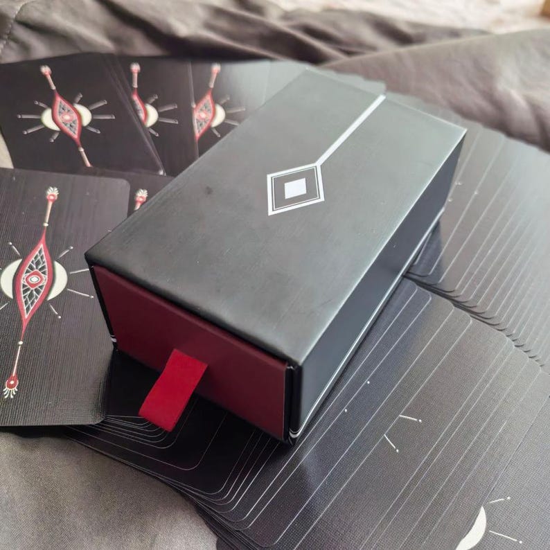 Puede incluir: Una caja rectangular negra con interior rojo y un dise&ntilde;o de rombo blanco en la parte superior. La caja est&aacute; parcialmente abierta, revelando una leng&uuml;eta roja. Varias cartas negras con dise&ntilde;os blancos y rojos son visibles.