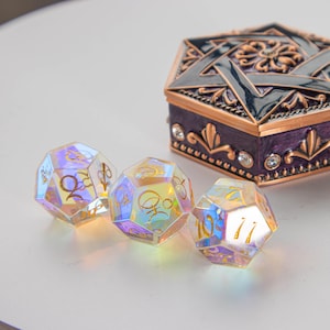 以下が含まれることがあります： 金色の刻印が施されたシンボルと数字を持つ、虹色に輝く多面体のサイコロ3個。その隣には、複雑なフィリグリー細工と小さな透明なクリスタルが埋め込まれた、濃い紫色と銅色の六角形の金属製ボックスが置かれています。