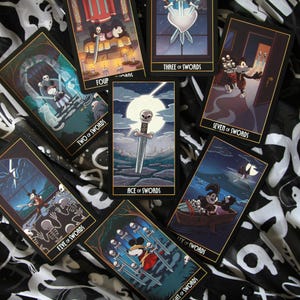 May include: A collection of tarot cards featuring illustrations with a cartoon theme. The cards display various scenes with swords, skulls, and other symbolic imagery. The cards are arranged on a black and white patterned surface.