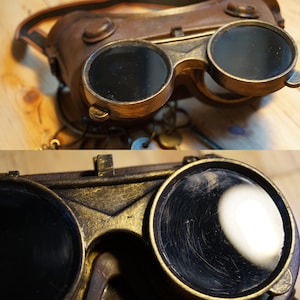 May include: A pair of steampunk-style goggles with round, dark lenses and a gold-colored metal frame. The goggles have a leather strap and are made of metal.