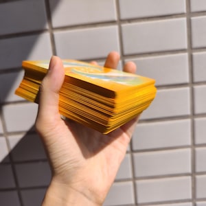 May include: A deck of tarot cards with yellow edges and a yellow cover is held in a hand. The cards are fanned out, revealing the edges. The background is a white tiled wall, with sunlight casting shadows.