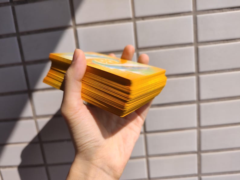 May include: A deck of tarot cards with yellow edges and a yellow cover is held in a hand. The cards are fanned out, revealing the edges. The background is a white tiled wall, with sunlight casting shadows.