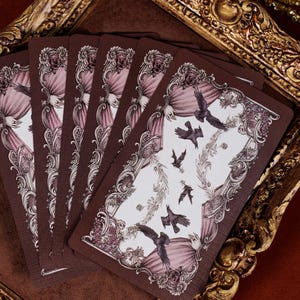 May include: A set of rectangular playing cards featuring a dark brown border with ornate floral and scrollwork patterns. The central white area displays illustrations of black birds and bats flying amidst delicate vine-like floral wreaths.