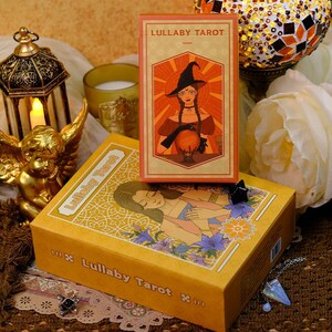 K&ouml;nnte beinhalten: Ein Tarotkartenspiel und ein Karton-Set mit dem Titel "Lullaby Tarot". Die gelbe Box zeigt eine Illustration einer Frau und Blumen. Das Kartenspiel zeigt eine Hexe mit einer Kristallkugel. Der Hintergrund umfasst Kerzen und dekorative Gegenst&auml;nde.