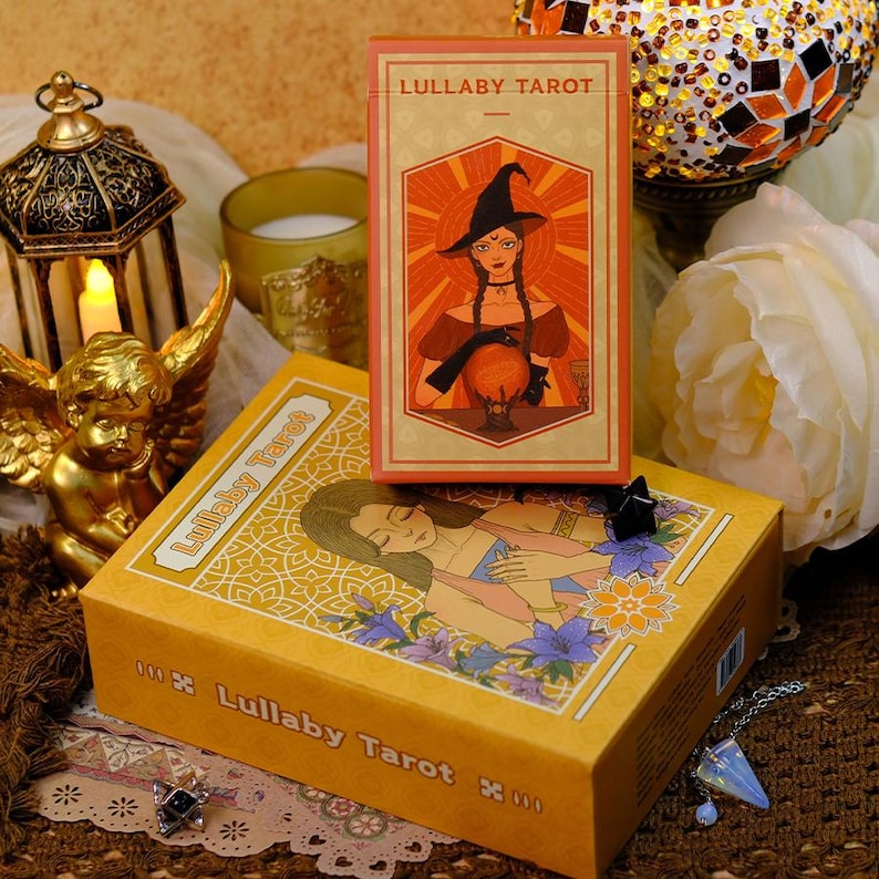 K&ouml;nnte beinhalten: Ein Tarotkartenspiel und ein Karton-Set mit dem Titel "Lullaby Tarot". Die gelbe Box zeigt eine Illustration einer Frau und Blumen. Das Kartenspiel zeigt eine Hexe mit einer Kristallkugel. Der Hintergrund umfasst Kerzen und dekorative Gegenst&auml;nde.