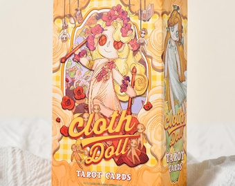 May include: A tarot card box with a whimsical illustration of a cloth doll character. The box is predominantly yellow with a checkered pattern and features the words "Cloth Doll Tarot Cards" in a playful font. The artwork includes floral and doll-like imagery.